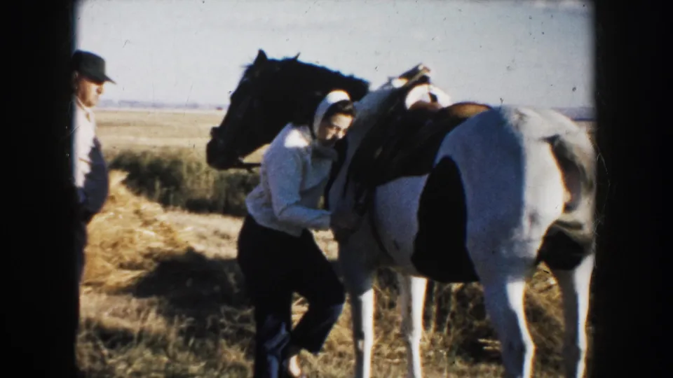 Woman in headscarf stands by a saddled black and white pinto horse as a man in a cap watches in a rural field.