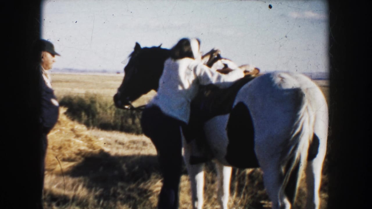 Woman climbs onto a black-and-white horse while a man in a cap stands nearby in an open field.