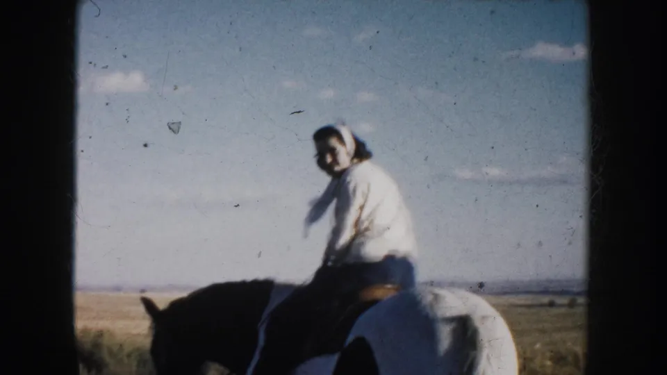 1953 South Dakota frame of a woman in a light blouse and headscarf riding a black-and-white horse through open prairie.