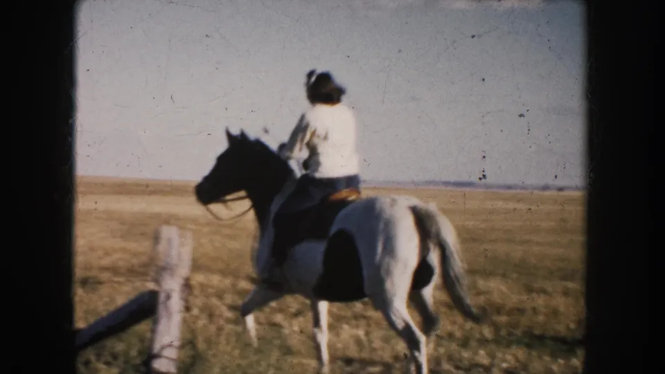 In 1953 South Dakota home-movie footage, a woman rides a pinto horse away from the camera across an open field under a pale sky.