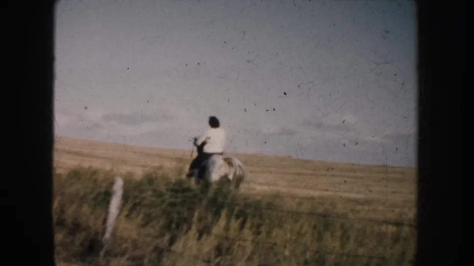 The rider appears smaller in the frame as the horse moves beyond taller grass and fence posts in the rural South Dakota landscape.