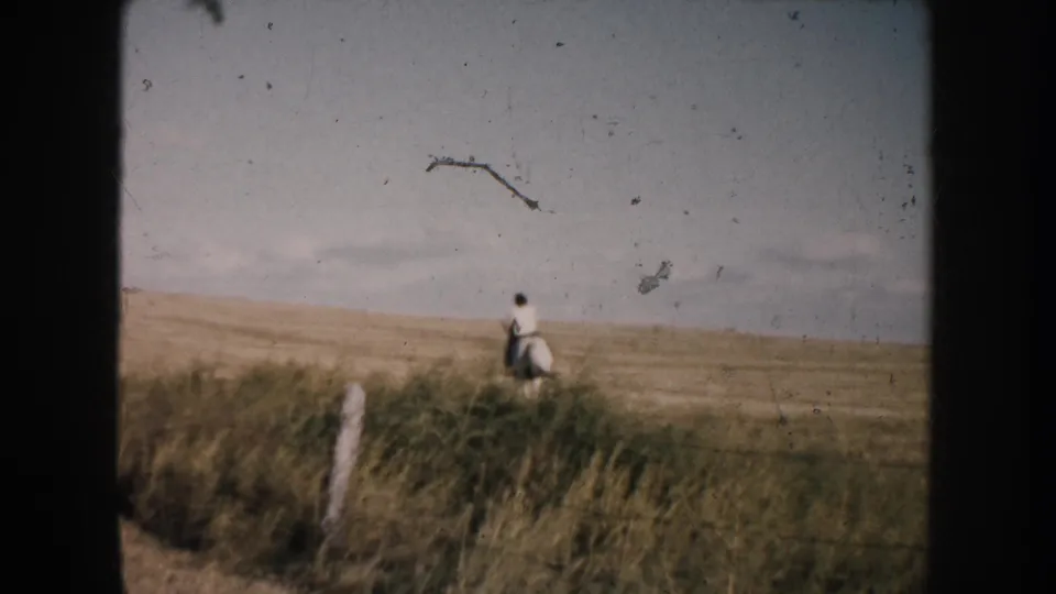 A woman on horseback appears small against the South Dakota prairie, with dry grass and a fence post visible in the foreground.
