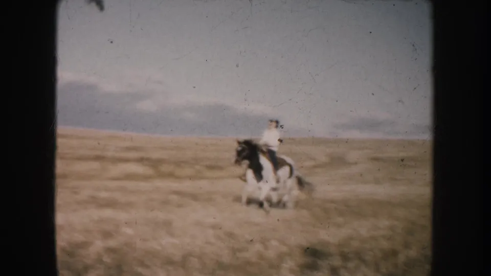The black-and-white horse carries the rider across South Dakota grassland, with soft 8mm blur and faded color.