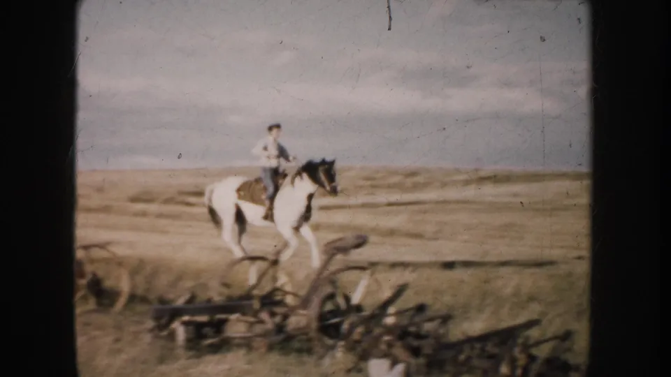 The rider and pinto horse move left to right past low farm machinery in the 1953 South Dakota landscape.