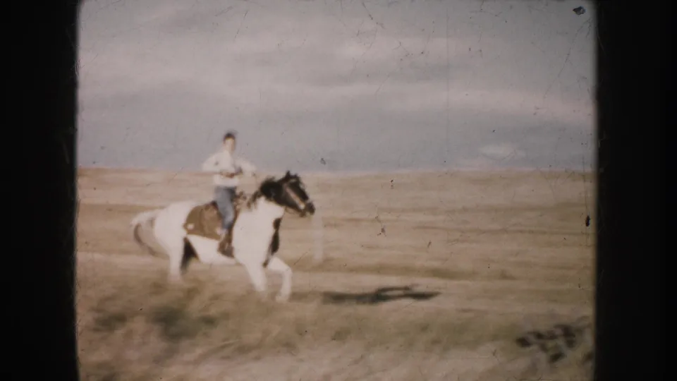 A rider in blue pants rides a black-and-white pinto horse across dry grass in South Dakota, 1953.