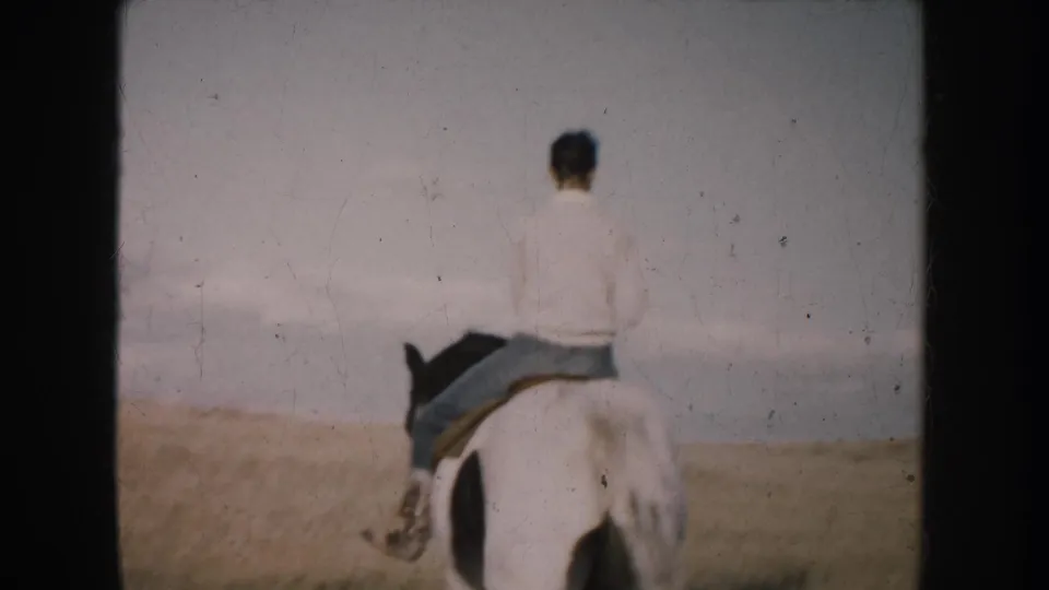 1953 South Dakota 8mm frame of a rider from behind on a black and white horse crossing pale prairie grass.