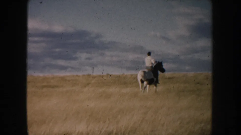 The rider continues away through tall prairie grass, with the pinto horse partly softened by 8mm blur and distance.