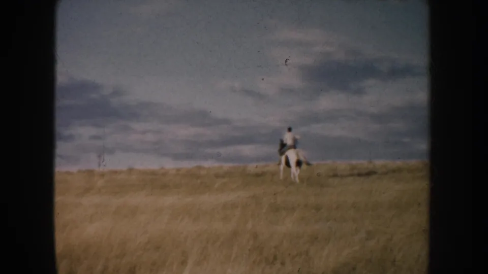 The lone rider on a white horse is centered against a wide 1950s South Dakota prairie view with a cloudy sky and soft 8mm color.