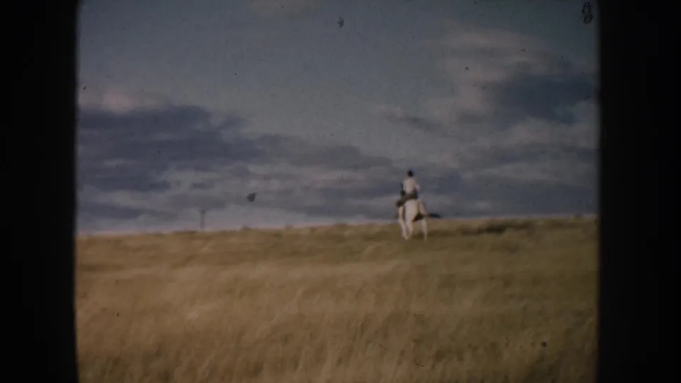 A distant rider on a white horse travels along the South Dakota prairie horizon, with tall grass in the foreground and heavy clouds above.