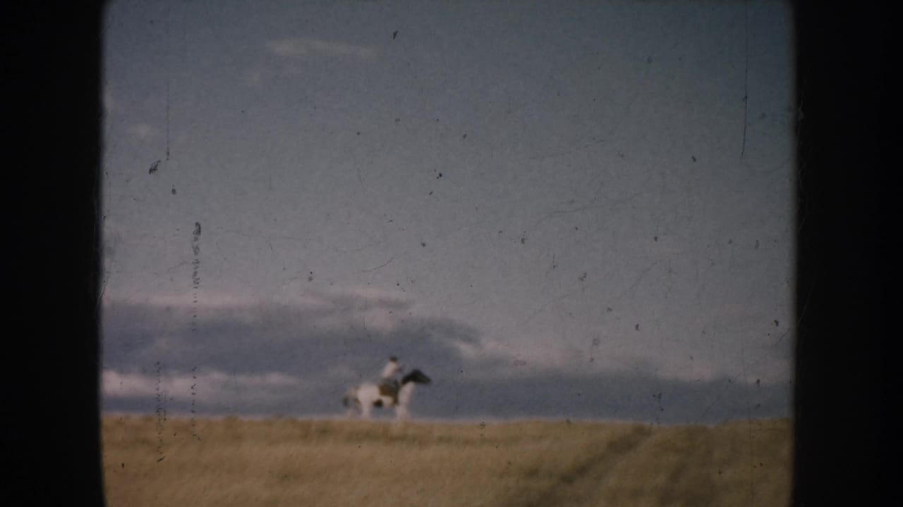 Small rider on a horse moving across a grassy rise under a muted sky.