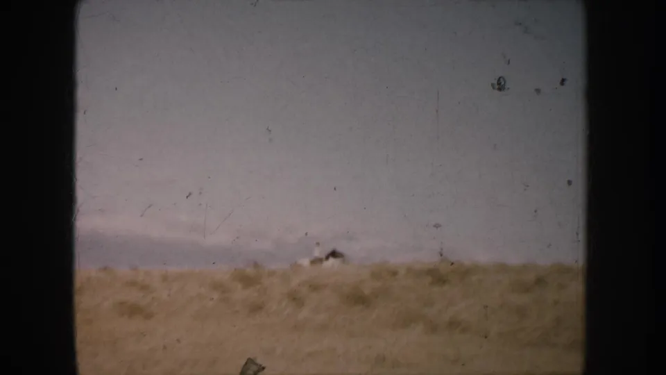 1953 home-movie frame with a distant horse and rider dropping behind sandy ground in South Dakota open country.