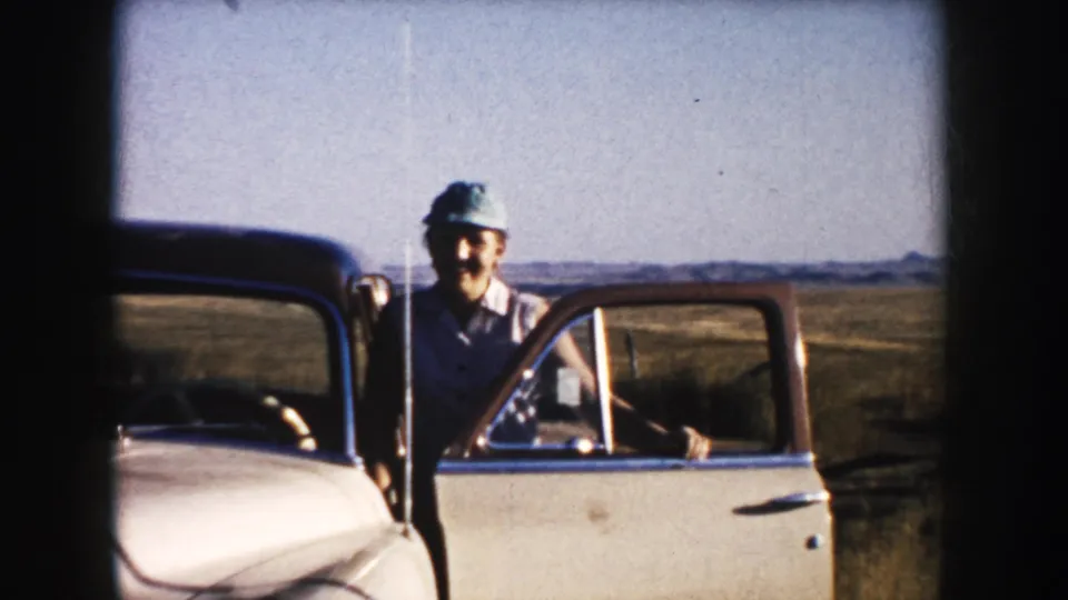 In South Dakota 1953 home-movie footage, a smiling man stands beside an older car with open doors against fields and distant hills.