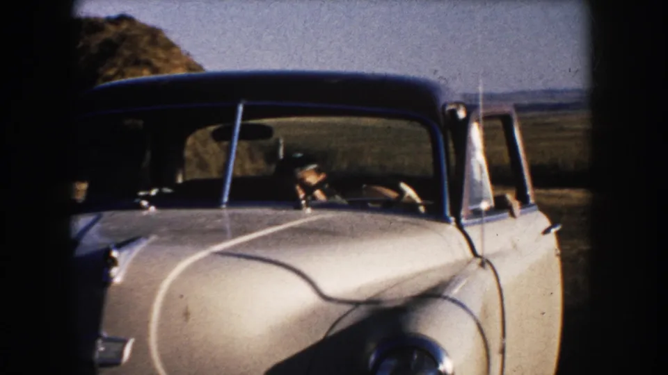 1953 South Dakota home-movie frame showing a white rounded car with a hat-wearing driver on a rural road near open fields.