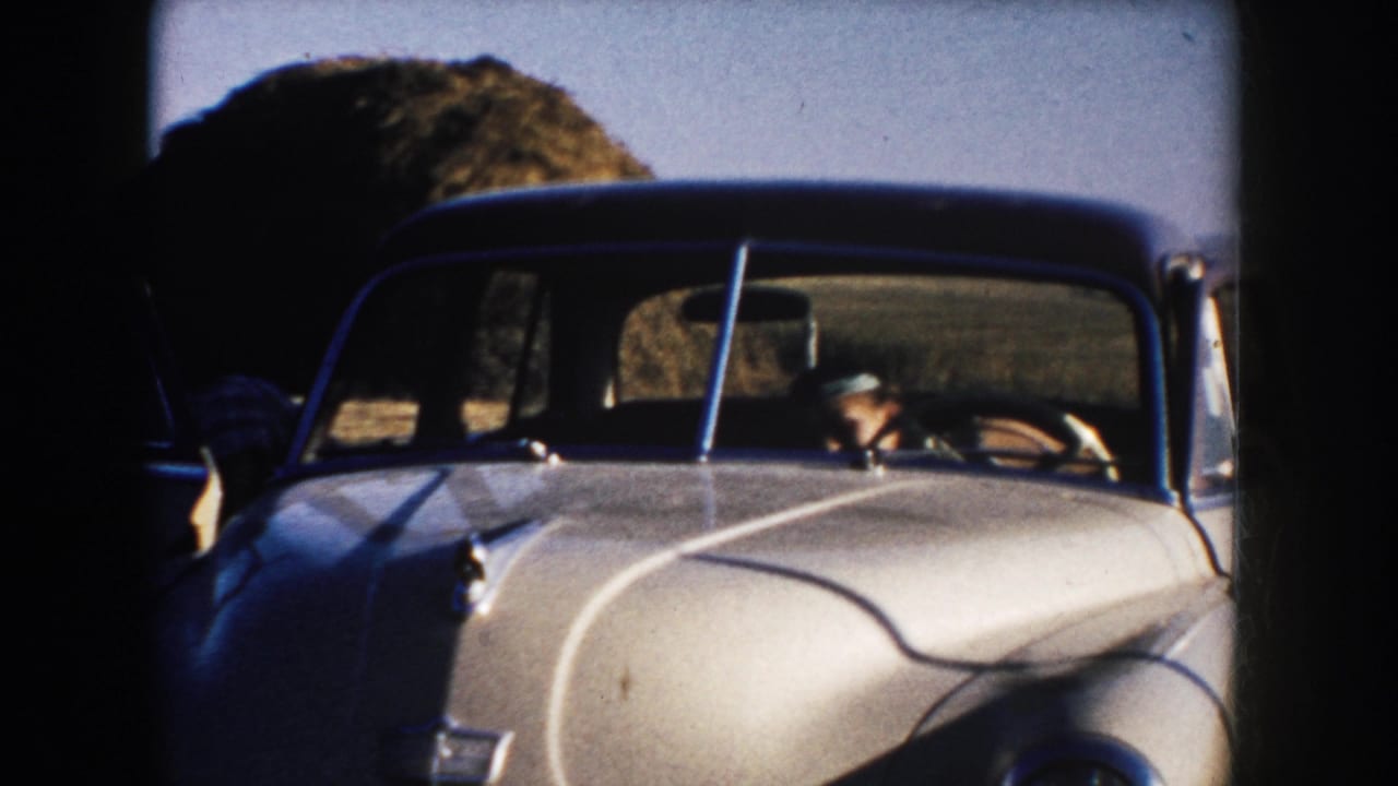 A white mid-century car passes through rural South Dakota farmland, with the driver visible behind the windshield.