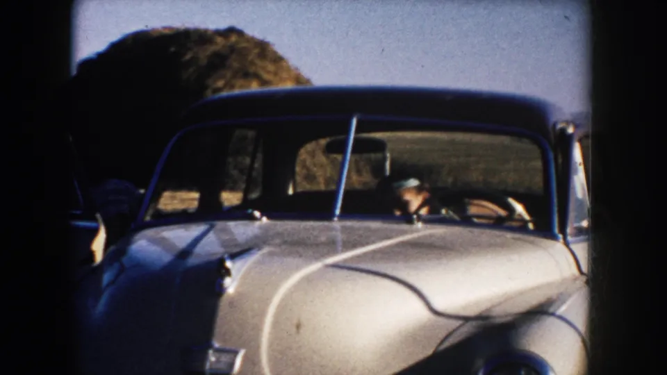 A white mid-century car passes through rural South Dakota farmland, with the driver visible behind the windshield.