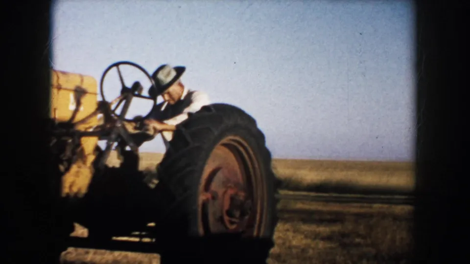 1953 rural South Dakota frame showing a man operating a yellow tractor with large rear tire in open farmland.