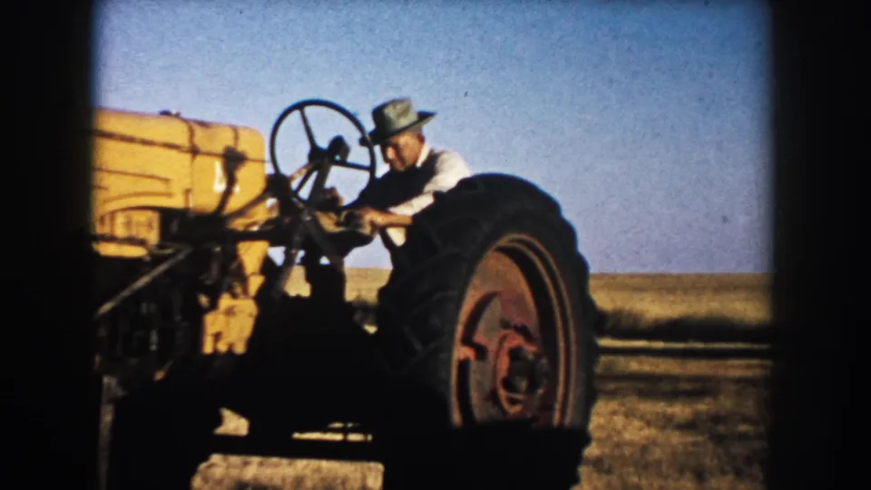 A man works at the controls of a yellow tractor in open South Dakota farmland, filmed in 1953 home-movie footage.