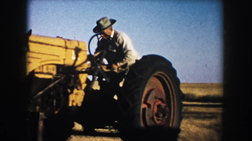 In 1953 South Dakota footage, a farmer in a light shirt and brimmed hat climbs onto a yellow tractor under a clear sky.
