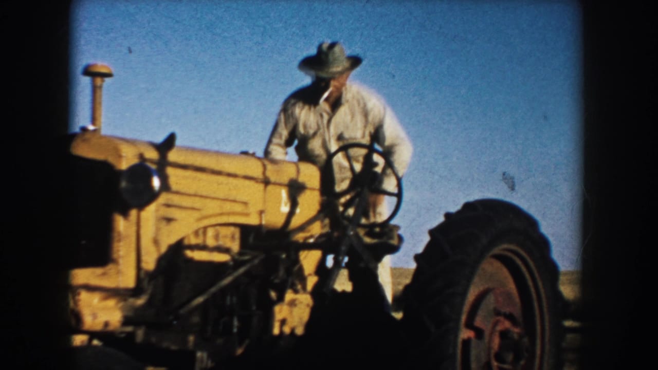 Farmer seated at the steering wheel of a yellow tractor in a flat field.