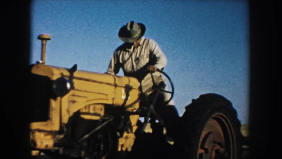 The 1953 South Dakota home-movie frame shows the farmer leaning toward the controls of a yellow tractor in a bare field.