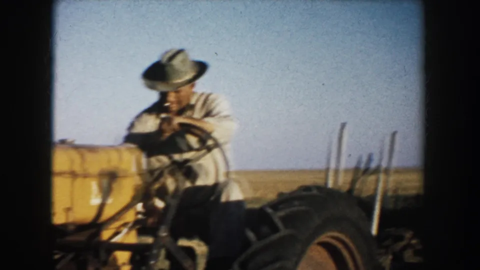A closer 8mm frame shows the farmer operating or preparing the yellow tractor in a South Dakota field, with the rear wheel prominent.