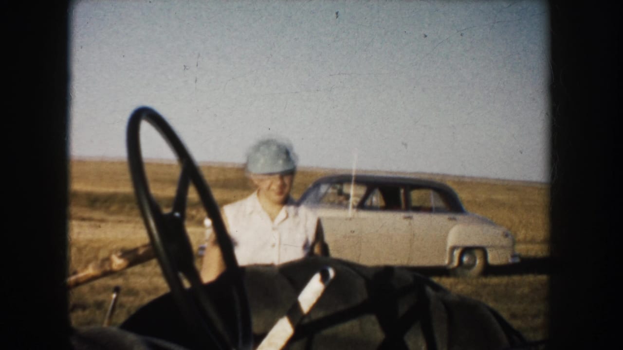 In 1953 South Dakota home-movie footage, a young woman in a cap stands by tractor controls with a rounded sedan parked in the rural field behind her.