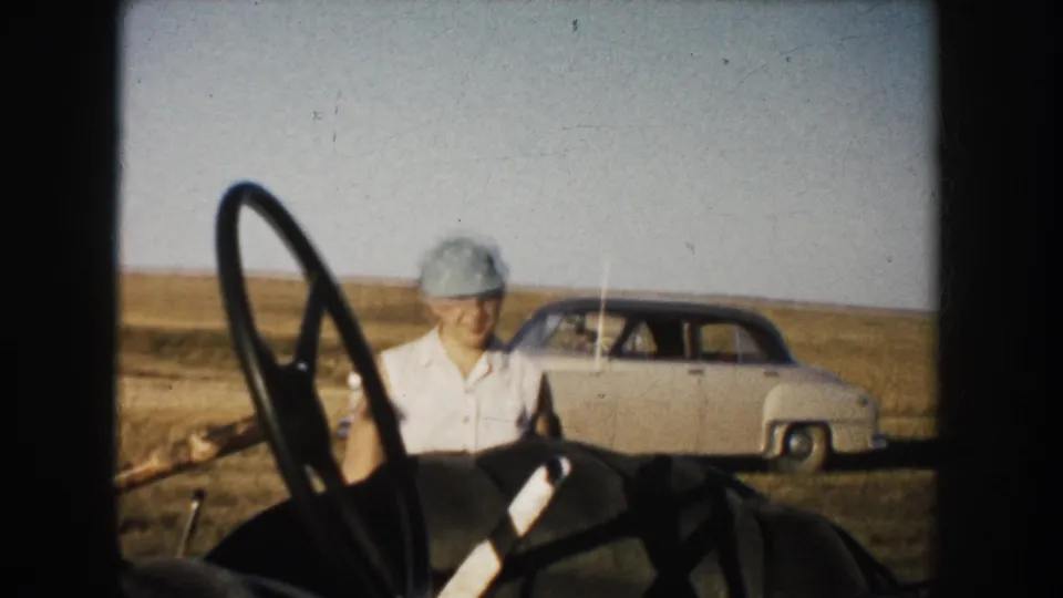 In 1953 South Dakota home-movie footage, a young woman in a cap stands by tractor controls with a rounded sedan parked in the rural field behind her.