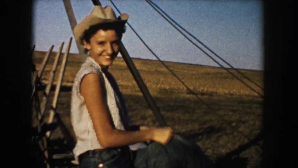 A smiling young woman in a cowboy hat sits near farm equipment and a wire fence in the South Dakota countryside.