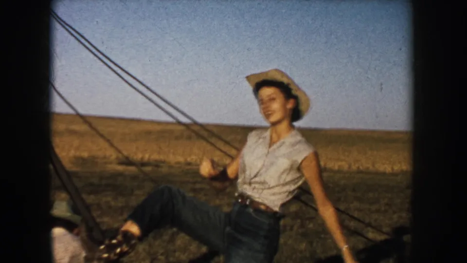 A young woman in a cowboy hat leans playfully near wire fencing in a South Dakota field, with diagonal fence lines crossing the frame.