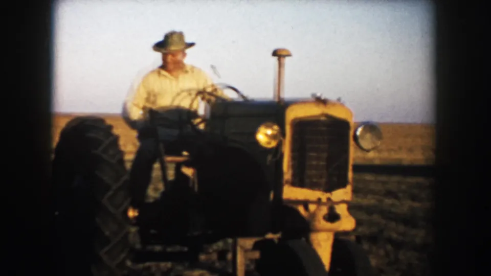 The farmer drives the tractor close to the camera, with the front grille and large wheel prominent in the 1953 farm scene.