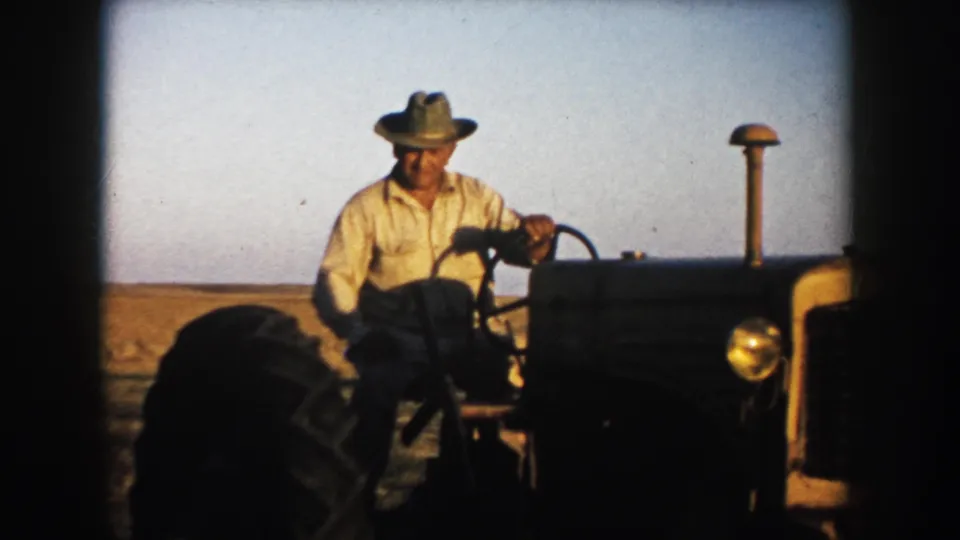 A farmer sits at the wheel of a tractor beside open South Dakota farmland, with warm 8mm color and soft exposure.
