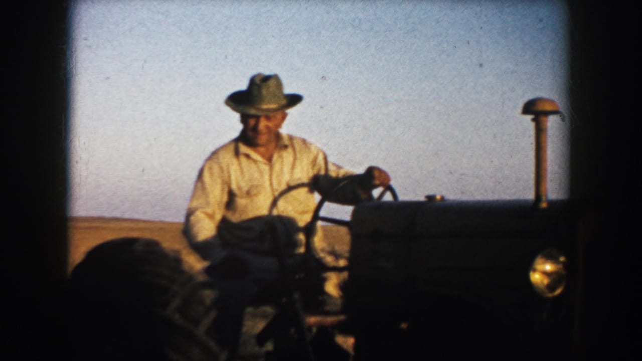 Farmer in a hat drives a tractor across an open field under a clear sky.