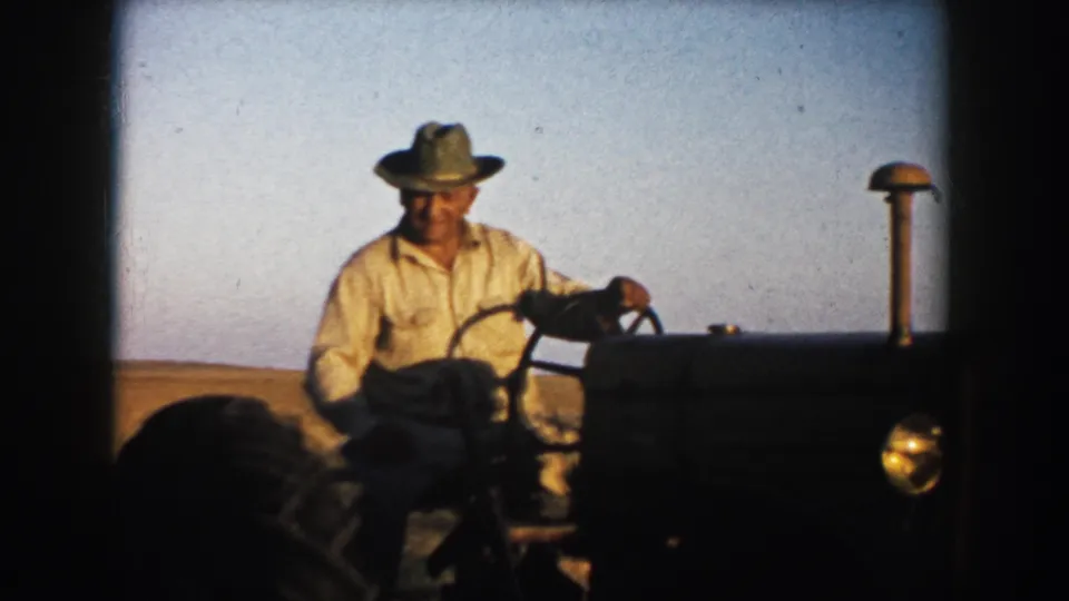 Adult farmer in South Dakota, 1953 metadata, steering a tractor through open farmland in warm light.