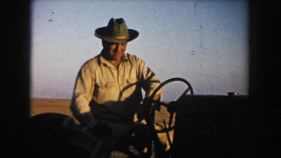 Close view of a 1953 South Dakota farmer operating a tractor, with broad field and clear sky in the background.