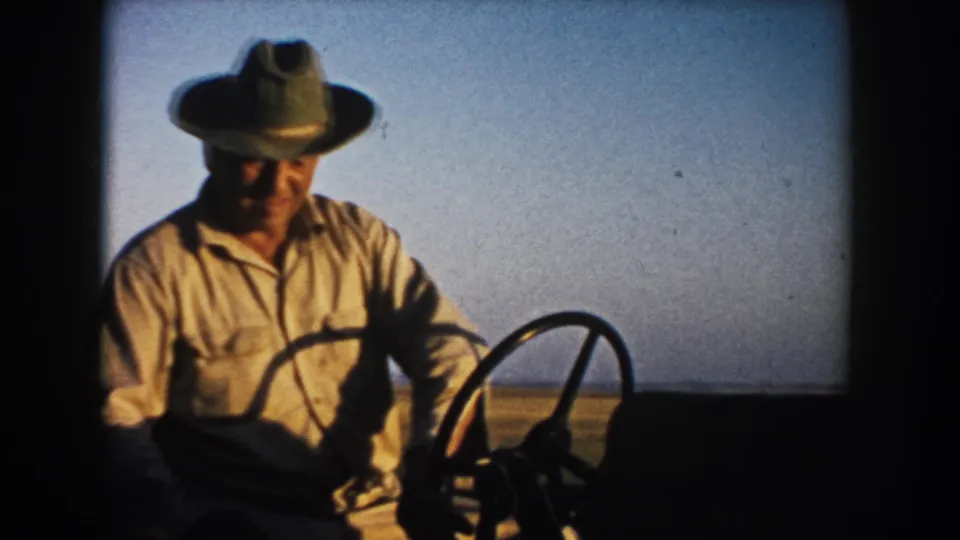 Adult farmer in hat and work shirt drives a tractor in South Dakota farmland, with 1953 date supported by metadata.