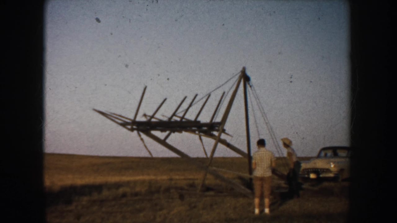 Two men stand near a large wooden frame in an open field with a car parked nearby.