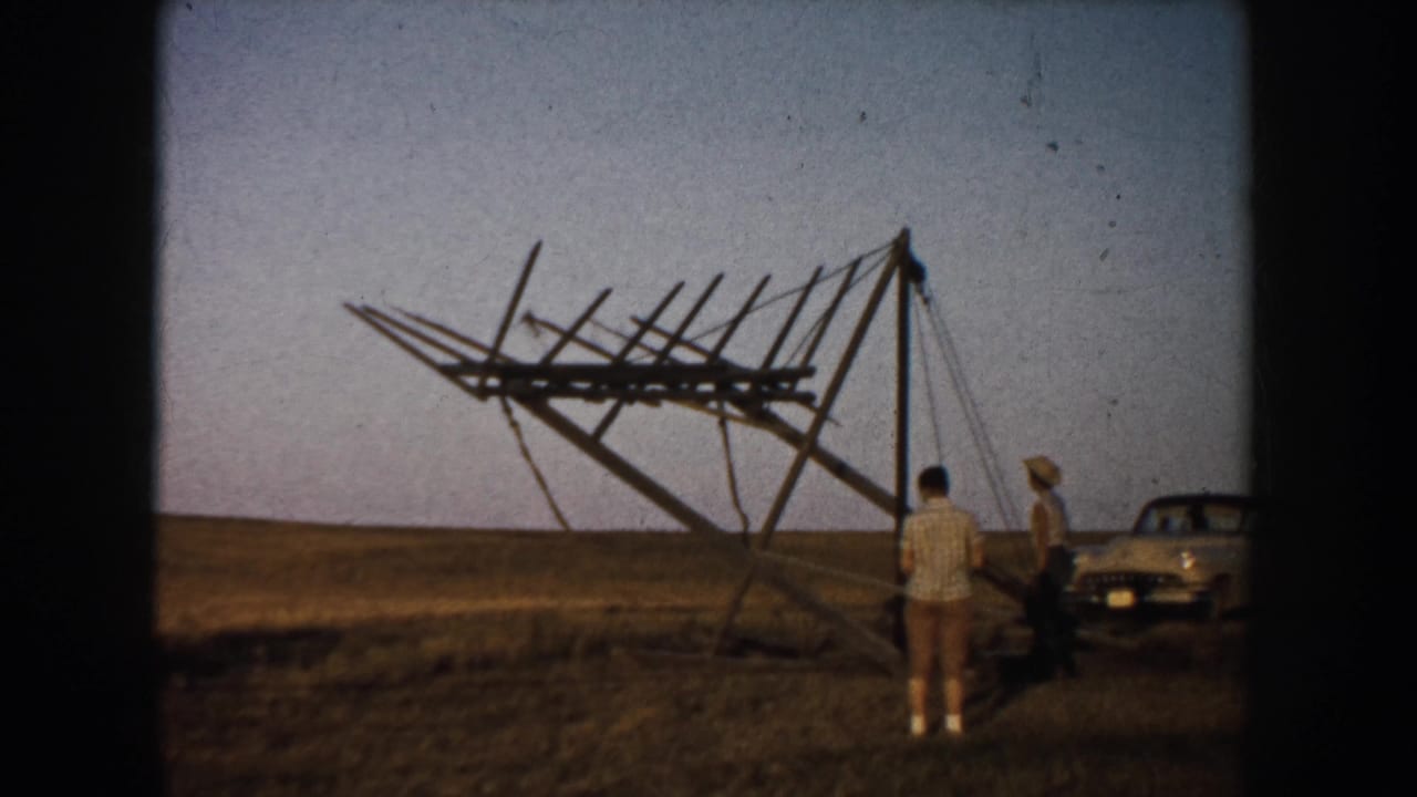 The sign-like wooden structure is partly raised in a South Dakota field as men stand near it and a 1950s car remains parked nearby.