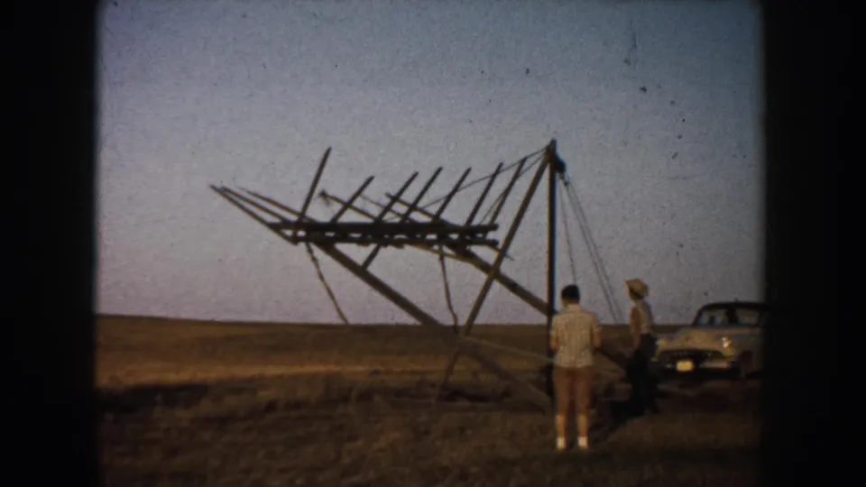 The sign-like wooden structure is partly raised in a South Dakota field as men stand near it and a 1950s car remains parked nearby.