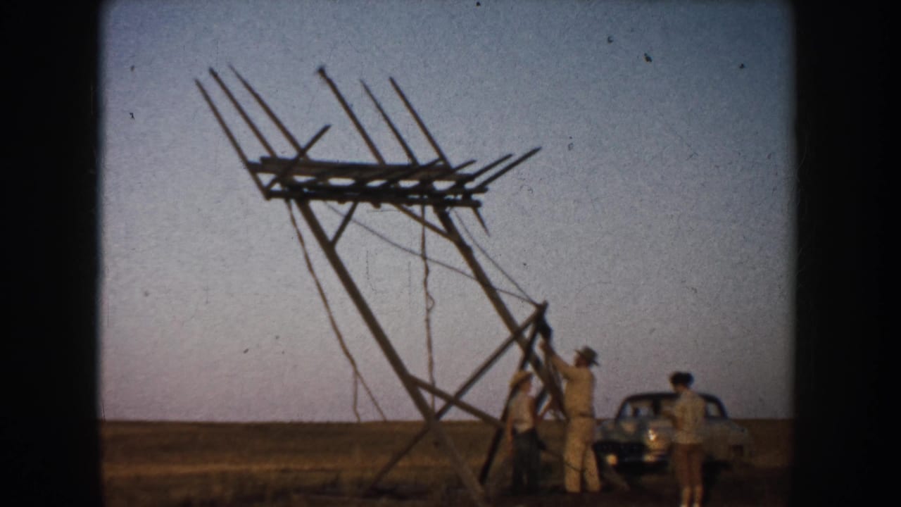 The large wooden frame leans high above the field while men gather at its base and a mid-century car sits close by.