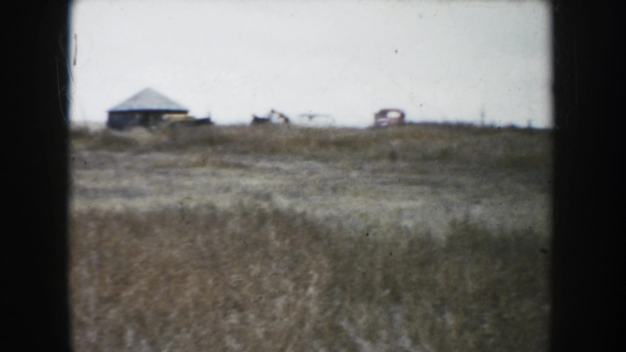 A distant view across a South Dakota field shows low farm buildings and vehicles on the horizon in faded 8mm color.