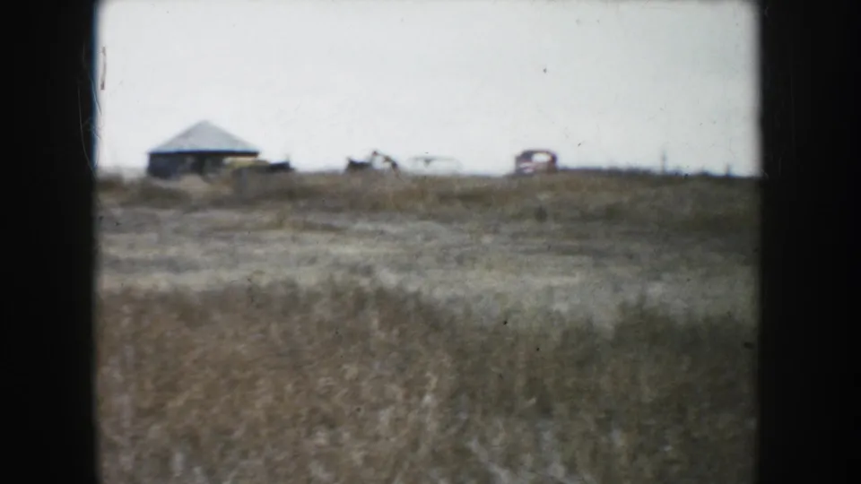 A distant view across a South Dakota field shows low farm buildings and vehicles on the horizon in faded 8mm color.