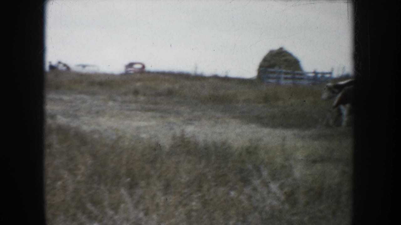 1953 South Dakota 8mm frame showing dry grassland, distant fencing, a haystack or hay bale, a small red car, and cattle at the edge of the pasture.