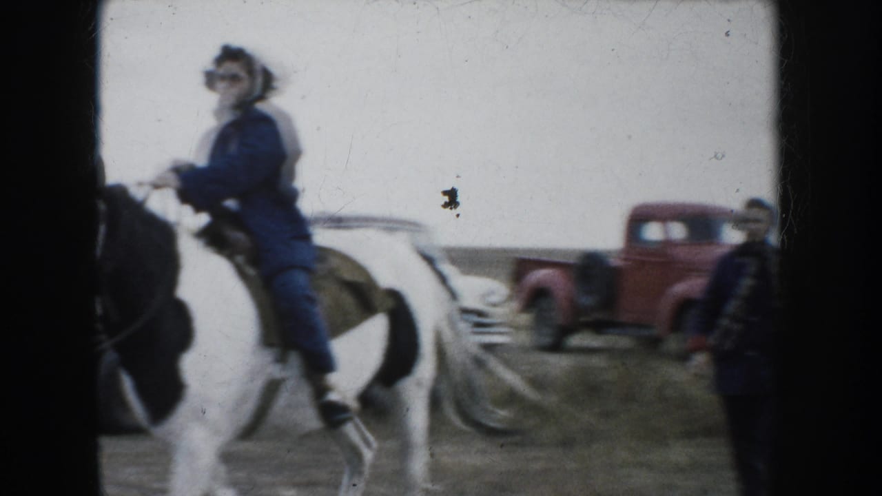 A woman in a blue coat rides a black-and-white pinto horse near a red pickup truck in rural South Dakota.