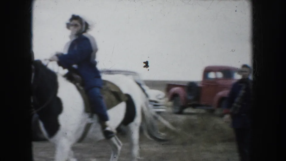 A woman in a blue coat rides a black-and-white pinto horse near a red pickup truck in rural South Dakota.