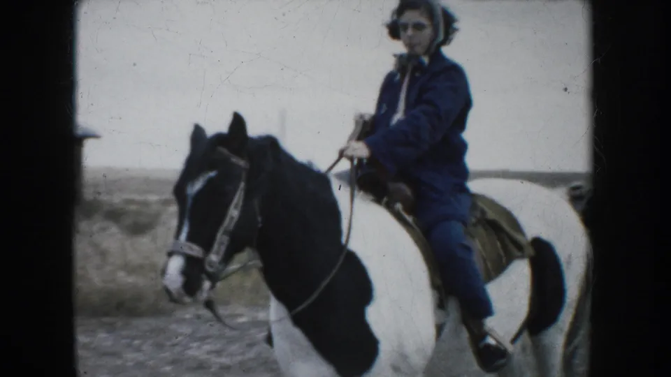 A woman wearing a blue coat and scarf rides a black-and-white pinto horse toward the camera in a 1953 South Dakota home movie.