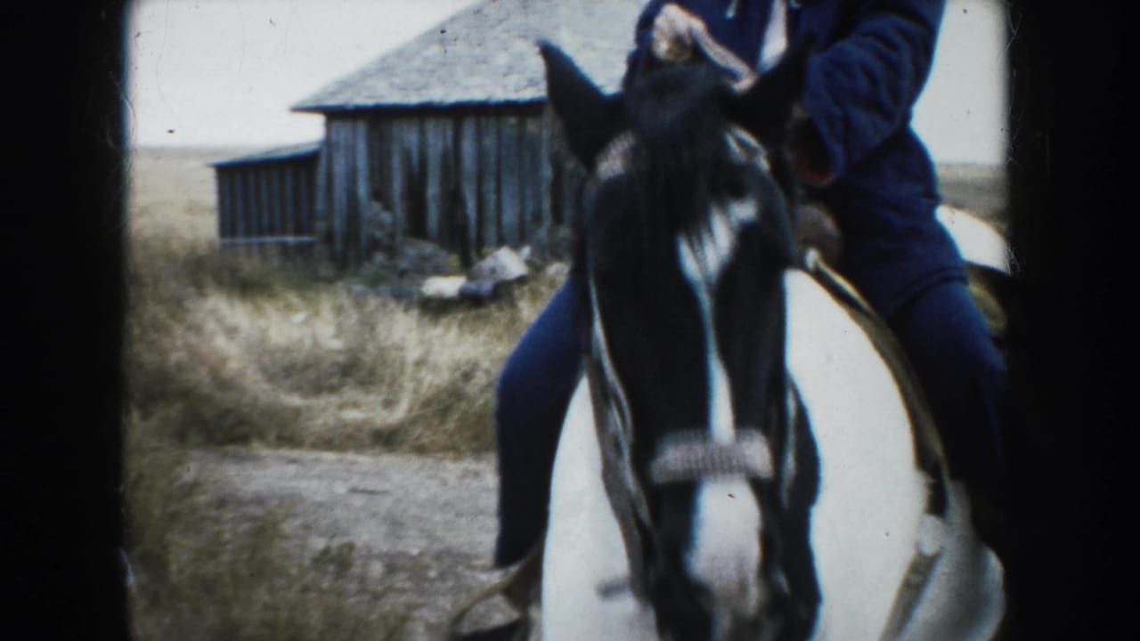 The pinto horse and rider pass close to the camera with a weathered wooden barn visible behind them in rural South Dakota.