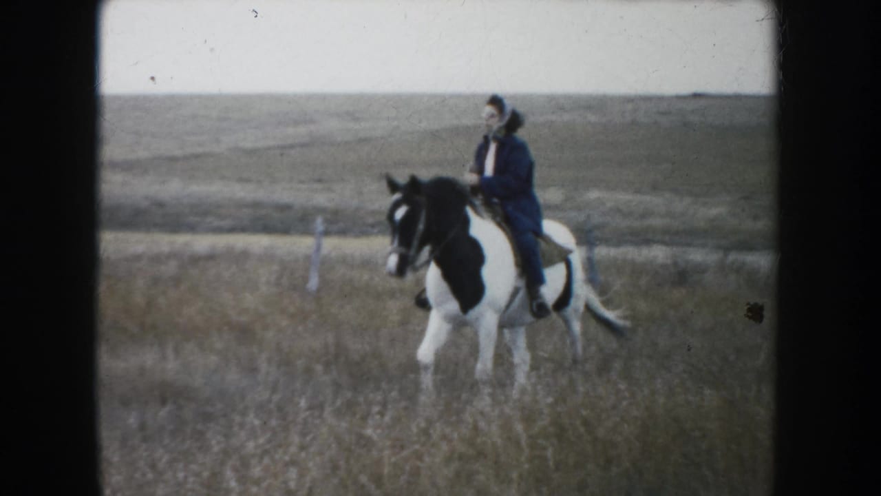 1953 South Dakota 8mm frame of a woman in a dark coat and hat riding a pinto horse across open prairie.