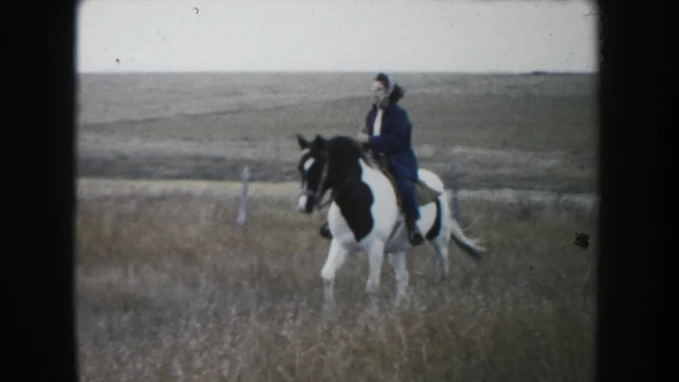 1953 South Dakota 8mm frame of a woman in a dark coat and hat riding a pinto horse across open prairie.