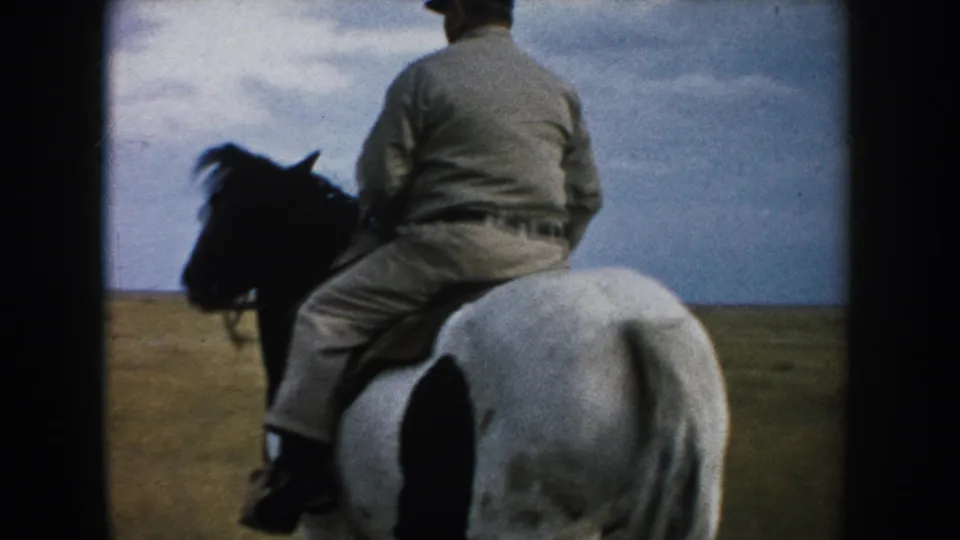 An older rider in light clothing rides away on a white horse with dark markings across open South Dakota prairie.