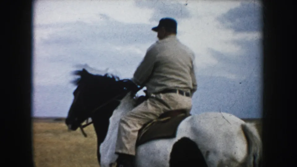 Adult man in work clothes and cap rides a brown-and-white horse across South Dakota grassland in 1953 home-movie footage.